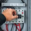 A hand adjusting a changeover switch in an electrical panel with visible wires and circuit breakers. Title